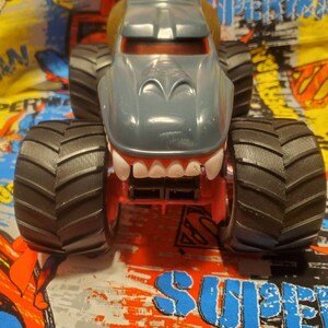 May include: A gray monster truck toy with a shark-like design. The truck has large black tires, a red chassis, and a wide, toothy grin. The cab is gray with a brown top. The background is a colorful Superman-themed fabric.