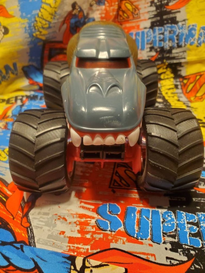 May include: A gray monster truck toy with a shark-like design. The truck has large black tires, a red chassis, and a wide, toothy grin. The cab is gray with a brown top. The background is a colorful Superman-themed fabric.