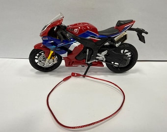 May include: A red, blue, and white Honda CBR motorcycle model with black tires and a black seat. A red cord is in the foreground. The motorcycle has a clear windshield and gold-colored front forks.