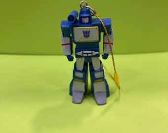 May include: A blue and white robot toy with a gold string for hanging. The robot has a purple emblem on its chest and is standing on a green surface. The robot is a toy from the Transformers franchise.