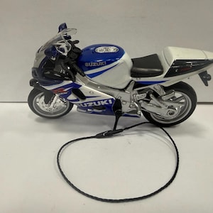Op de afbeelding: Gedetailleerd model van een Suzuki-motorfiets, in blauw en wit met zilveren details. De miniatuurmotorfiets heeft realistische kenmerken, waaronder wielen, een zadel en het Suzuki-logo. Een zwart koord staat op de voorgrond.