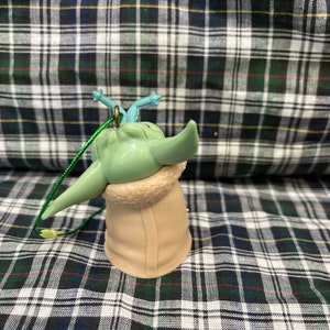 May include: A green and blue plastic bauble featuring a character from the Star Wars franchise, wearing a brown coat. The bauble has a green string attached to the top.