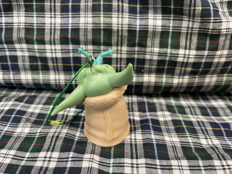 May include: A green and blue plastic bauble featuring a character from the Star Wars franchise, wearing a brown coat. The bauble has a green string attached to the top.