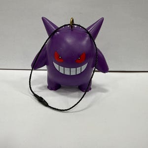 May include: A purple Gengar figurine with red eyes and a wide, toothy grin. The figure has pointed ears and small arms and legs. A black cord is attached to the top. This collectible is a Pokemon character.