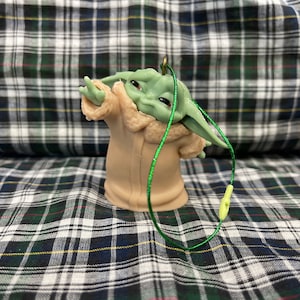 May include: A green and brown figurine of a character from the Star Wars franchise, wearing a brown coat with a fur collar. The figurine is attached to a green string.