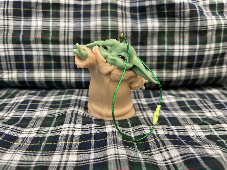 May include: A green and brown figurine of a character from the Star Wars franchise, wearing a brown coat with a fur collar. The figurine is attached to a green string.