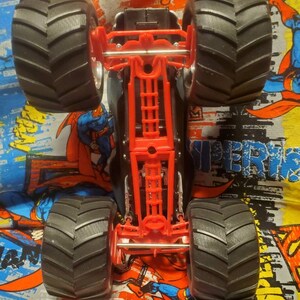 May include: A black and red monster truck toy with large, black, grooved tires. The truck's chassis is red, and the body is black. The image is taken from underneath the truck, showing the underside details.