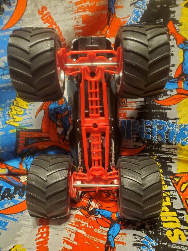 May include: A black and red monster truck toy with large, black, grooved tires. The truck's chassis is red, and the body is black. The image is taken from underneath the truck, showing the underside details.