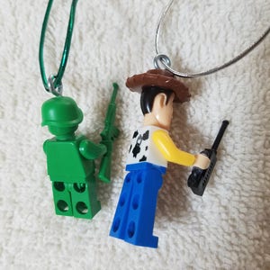 May include: Two toy figure necklaces. One is a green army man with a rifle and the other is a cowboy with a walkie talkie. Both figures are wearing necklaces.