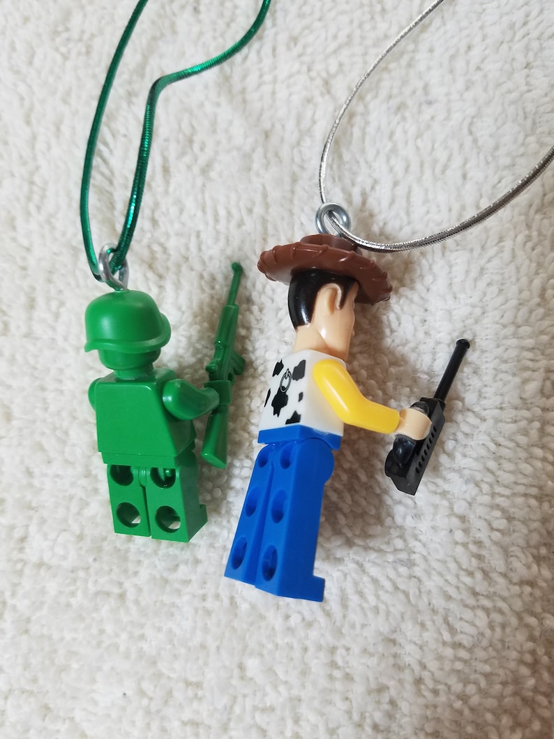 May include: Two toy figure necklaces. One is a green army man with a rifle and the other is a cowboy with a walkie talkie. Both figures are wearing necklaces.