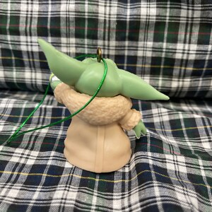 以下が含まれることがあります： スター・ウォーズのフランチャイズ、マンダロリアンのキャラクターの緑と茶色のオーナメント。キャラクターは、ファーのトリムが付いた茶色のコートを着ており、大きな耳をしています。オーナメントには緑色の紐が付いています。