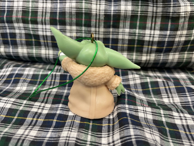 以下が含まれることがあります： スター・ウォーズのフランチャイズ、マンダロリアンのキャラクターの緑と茶色のオーナメント。キャラクターは、ファーのトリムが付いた茶色のコートを着ており、大きな耳をしています。オーナメントには緑色の紐が付いています。