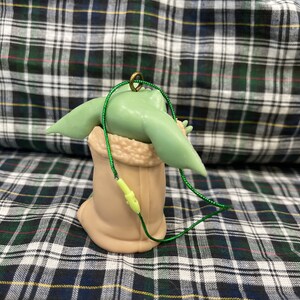 Pode incluir: Um enfeite de Natal verde e marrom com um personagem da franquia Star Wars, The Mandalorian. O enfeite est&aacute; pendurado em um cord&atilde;o verde.