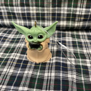 Peut inclure: Une d&eacute;coration de No&euml;l verte et marron en forme de personnage de la franchise Star Wars, The Mandalorian. L'ornement tient un petit objet noir dans ses mains.