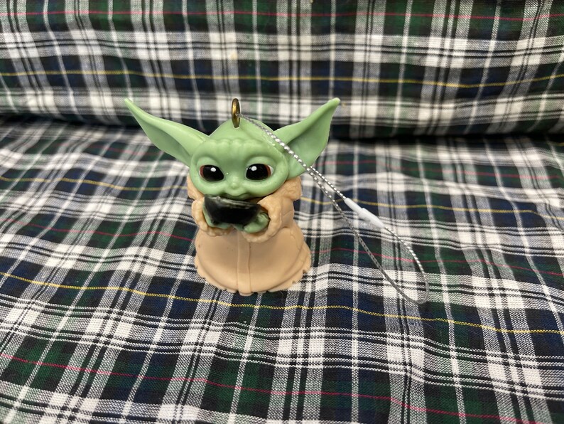 Peut inclure: Une d&eacute;coration de No&euml;l verte et marron en forme de personnage de la franchise Star Wars, The Mandalorian. L'ornement tient un petit objet noir dans ses mains.