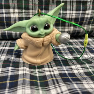 以下が含まれることがあります： スターウォーズのフランチャイズ、マンダロリアンのキャラクターの緑と茶色のプラスチック製のオーナメント。キャラクターは右手に銀色のボールを持っています。オーナメントには緑色の紐が付いています。