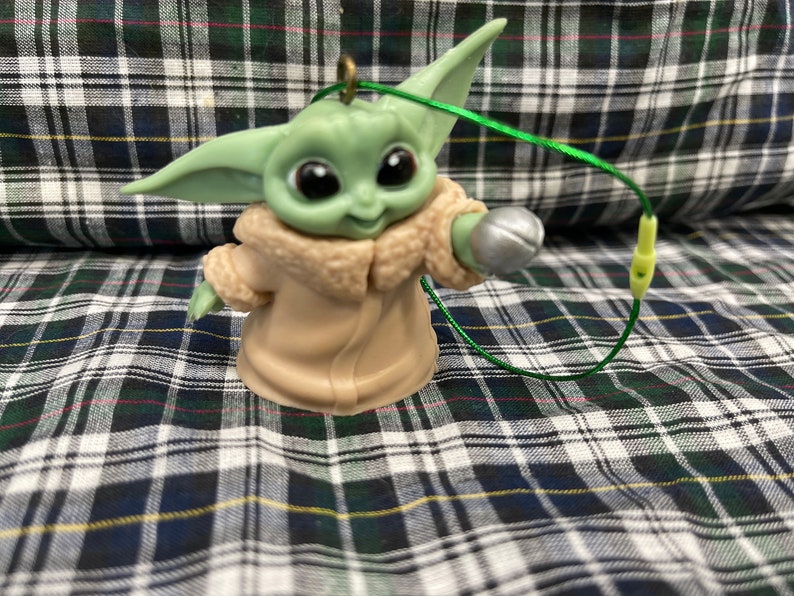 以下が含まれることがあります： スターウォーズのフランチャイズ、マンダロリアンのキャラクターの緑と茶色のプラスチック製のオーナメント。キャラクターは右手に銀色のボールを持っています。オーナメントには緑色の紐が付いています。