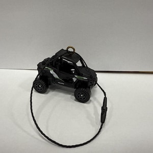 May include: Black Polaris toy UTV with green and white accents. The miniature off-road vehicle has large black tires and a black cord for hanging. A small gold-colored ring is attached to the top of the UTV.