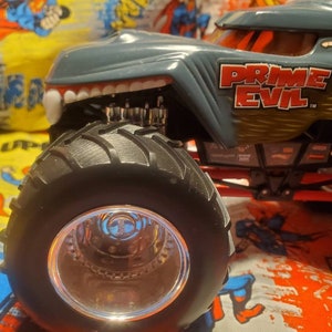 May include: A gray and gold toy monster truck with the text "PRIME EVIL" on the side. The truck has large black tires and a detailed design, including a toothy grill and a detailed engine.