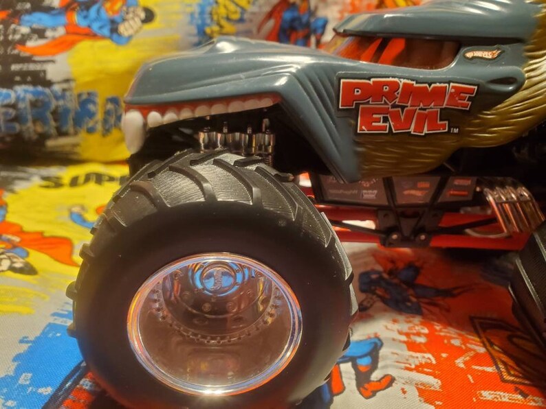 May include: A gray and gold toy monster truck with the text "PRIME EVIL" on the side. The truck has large black tires and a detailed design, including a toothy grill and a detailed engine.