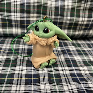 Pode incluir: Um enfeite de pl&aacute;stico verde e marrom de um personagem da franquia Star Wars, The Mandalorian. O personagem est&aacute; vestindo uma t&uacute;nica marrom e tem orelhas grandes. O enfeite tem uma corda verde amarrada.