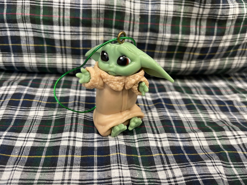 Pode incluir: Um enfeite de pl&aacute;stico verde e marrom de um personagem da franquia Star Wars, The Mandalorian. O personagem est&aacute; vestindo uma t&uacute;nica marrom e tem orelhas grandes. O enfeite tem uma corda verde amarrada.