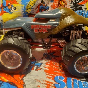 May include: A gray and brown Hot Wheels monster truck toy with the text "PRIME EVIL" on the side. The truck has large black tires with silver rims and a red chassis. The truck's design resembles a monster with sharp teeth.