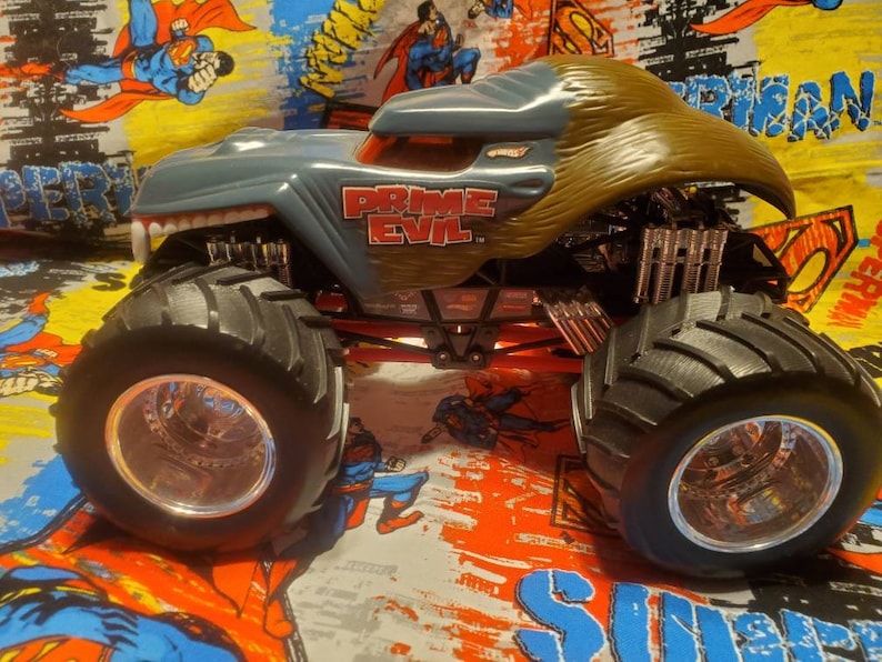 May include: A gray and brown Hot Wheels monster truck toy with the text "PRIME EVIL" on the side. The truck has large black tires with silver rims and a red chassis. The truck's design resembles a monster with sharp teeth.