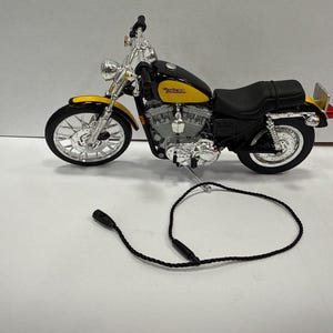May include: A detailed model motorcycle in black and yellow, featuring chrome accents on the wheels, engine, and handlebars. The motorcycle has a black seat and a black cord attached. The model is a replica of a Harley Davidson motorcycle.