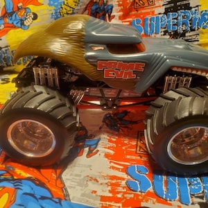 May include: A gray and tan Hot Wheels Prime Evil monster truck toy. The truck has large black tires, a red chassis, and the words "PRIME EVIL" in red lettering. The truck is on a colorful Superman-themed background.