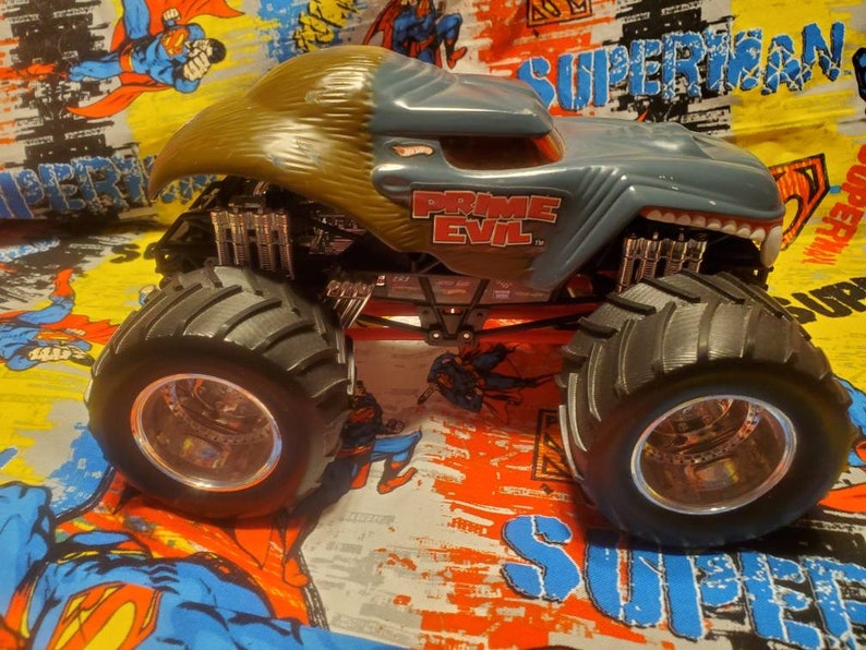 May include: A gray and tan Hot Wheels Prime Evil monster truck toy. The truck has large black tires, a red chassis, and the words "PRIME EVIL" in red lettering. The truck is on a colorful Superman-themed background.