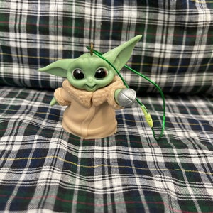 以下が含まれることがあります： スター・ウォーズのフランチャイズ、マンダロリアンのキャラクターの緑と茶色のプラスチック製のオーナメント。キャラクターは右手に銀色の物体を持ち、オーナメントには緑色の紐が付いています。