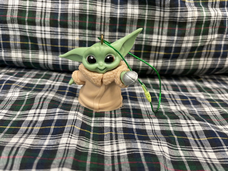 以下が含まれることがあります： スター・ウォーズのフランチャイズ、マンダロリアンのキャラクターの緑と茶色のプラスチック製のオーナメント。キャラクターは右手に銀色の物体を持ち、オーナメントには緑色の紐が付いています。