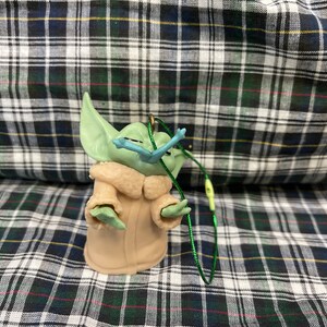 May include: A green and brown plush toy of a character from the Star Wars franchise, The Mandalorian. The toy is wearing a brown coat and has a green face with large ears. The toy is attached to a green string.