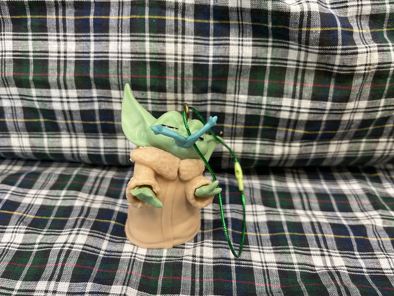 May include: A green and brown plush toy of a character from the Star Wars franchise, The Mandalorian. The toy is wearing a brown coat and has a green face with large ears. The toy is attached to a green string.