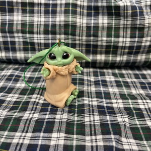 Pode incluir: Um ornamento de pel&uacute;cia verde e marrom de um personagem da franquia Star Wars, The Mandalorian. O personagem est&aacute; vestindo uma t&uacute;nica marrom e tem olhos grandes.