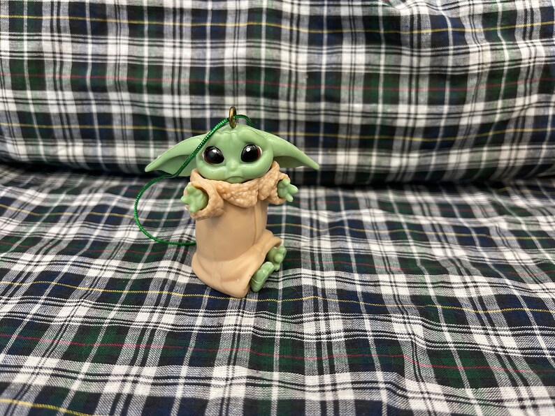 Pode incluir: Um ornamento de pel&uacute;cia verde e marrom de um personagem da franquia Star Wars, The Mandalorian. O personagem est&aacute; vestindo uma t&uacute;nica marrom e tem olhos grandes.