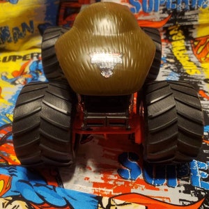 May include: A brown Monster Jam truck toy with large black tires. The truck has the Monster Jam logo on the front. The truck is red and black and sits on a colorful Superman-themed background.