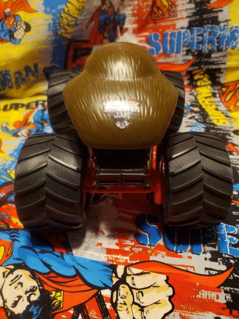 May include: A brown Monster Jam truck toy with large black tires. The truck has the Monster Jam logo on the front. The truck is red and black and sits on a colorful Superman-themed background.