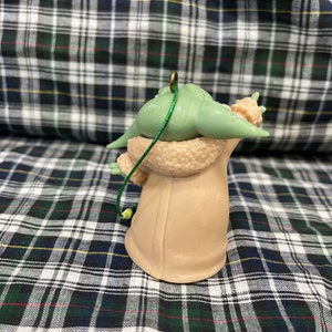 May include: A green and brown Christmas bauble shaped like a character from the Star Wars franchise. The bauble is wearing a brown coat and has a green string attached to the top.