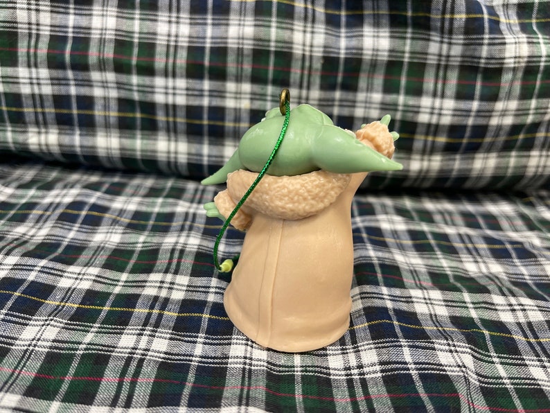 May include: A green and brown Christmas bauble shaped like a character from the Star Wars franchise. The bauble is wearing a brown coat and has a green string attached to the top.