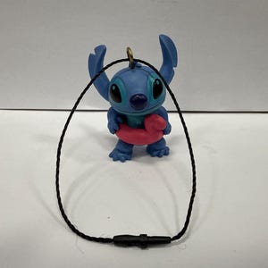 May include: A blue Stitch figurine with a pink inflatable ring, attached to a black cord necklace. The figurine has large ears and a friendly expression. The necklace has a black clasp.