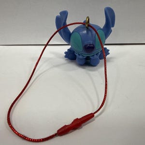 May include: A blue Stitch figurine with a red string attached. The figurine has large ears, a round nose, and teal accents. The red string has a red plastic clasp.