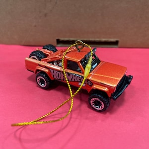 May include: Orange Hot Wheels toy truck with a black bumper and black wheels. The truck has a gold string attached to it, making it a Christmas ornament.