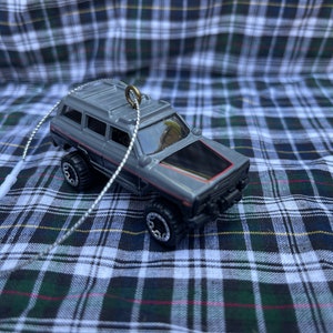 May include: A gray toy SUV with black and red accents, featuring large off-road tires. The vehicle has a black hood and a silver string for hanging. The background is a plaid pattern.
