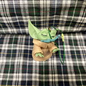 May include: A green and brown figurine of a character from the Star Wars franchise, wearing a brown coat and a blue moustache. The figurine is attached to a green string.