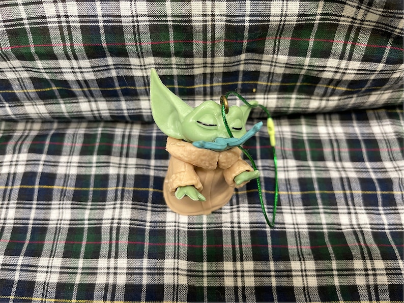 May include: A green and brown figurine of a character from the Star Wars franchise, wearing a brown coat and a blue moustache. The figurine is attached to a green string.