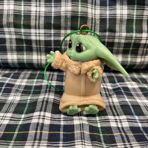 Pode incluir: Um enfeite de pl&aacute;stico verde e marrom de um personagem da s&eacute;rie Star Wars, The Mandalorian. O personagem est&aacute; vestindo um casaco marrom e tem olhos grandes.