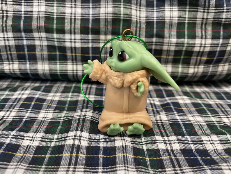Pode incluir: Um enfeite de pl&aacute;stico verde e marrom de um personagem da s&eacute;rie Star Wars, The Mandalorian. O personagem est&aacute; vestindo um casaco marrom e tem olhos grandes.