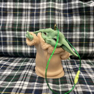 May include: A green and brown plastic bauble featuring a character from the Star Wars franchise, The Mandalorian. The character is wearing a brown coat with a fur trim and has large ears. The bauble has a green string attached to it.